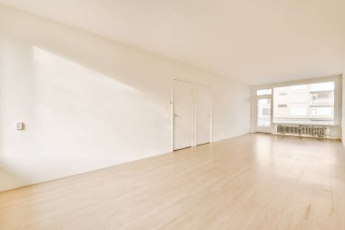 Odanın ortasında beyaz duvarları ve ahşap döşemeleri olan boş bir oda var. Sağ tarafta büyük bir pencere var.