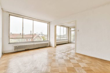 Boş bir oda, ahşap döşemeli ve geniş pencereli şehir merkezlerine bakan.