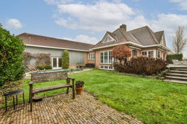 a house with a pool in the back yard and some bushes on the lawn, there is an empty bench