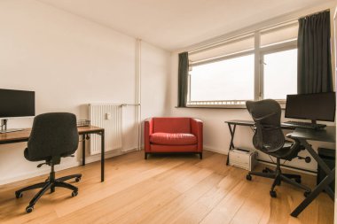 a living room with a red chair and computer on the desk in front of a large window that looks out onto the street