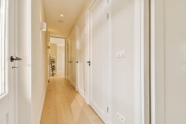 Beyaz duvarları ve ahşap döşemeleri olan uzun bir koridor pencerelerden gelen doğal ışıkla aydınlanır.