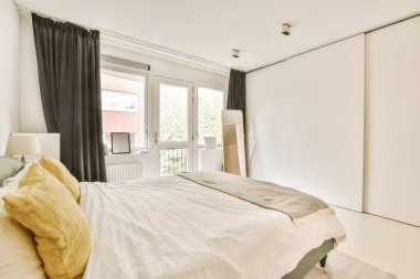 Beyaz duvarları ve siyah perdeleri olan bir yatak odası sokağa bakan açık bir pencerenin yanında.