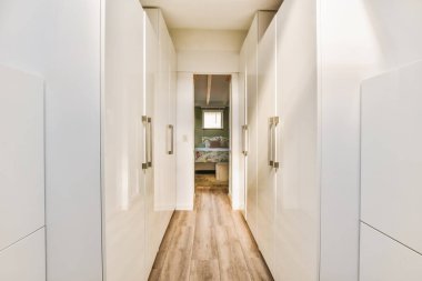 Beyaz duvarları ve ahşap zemini olan uzun dar bir koridor pencerelerden gelen doğal ışıkla aydınlanır.