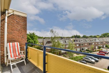 Kırmızı ve beyaz çizgili sandalyeleri, mavi gökyüzü ve yeşil ağaçları olan bir balkon bir tuğla duvardır.