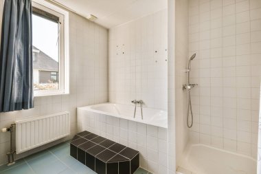 Duvarlarında mavi fayanslar ve beyaz fayanslar olan bir banyo. Bir de pencerenin yanında küvet var.