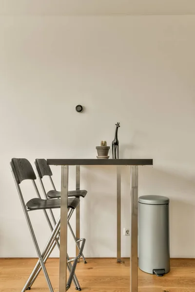 a table and chairs in a room with a trash can on the floor next to the table is a white wall