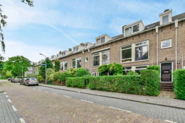 some houses on the side of a street with cars parked in front of them and bushes growing along the sidewalk
