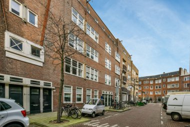 Amsterdam, Netherlands - 10 April, 2021: a city street with cars parked on the side and buildings in the back ground, there is a tree that has no leaves