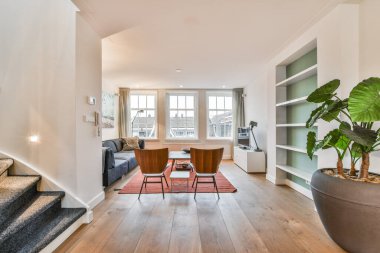 Odanın sol tarafında büyük bir bitki de dahil olmak üzere ahşap döşemeli ve beyaz duvarlı bir oturma odası.