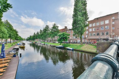 Amsterdam, Hollanda - 10 Nisan 2021: Bazı tekneler arka planda binalar ve nehir kıyısı boyunca uzanan yeşil ağaçlarla bir kanalın kenarına yanaştı
