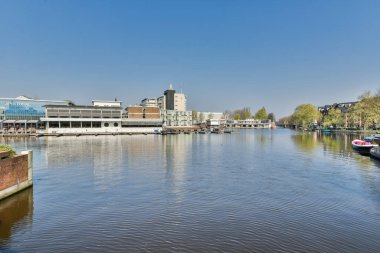 Amsterdam, Hollanda - 10 Nisan 2021: her iki yakasında tekneleri ve binaları olan bir nehir, kanal boyunca bir tekne turundan alındı.