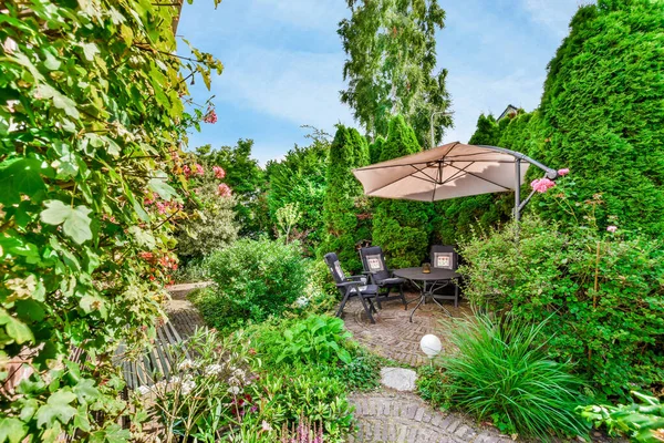 a patio with an umbrella and table in the middle of it, surrounded by lush green plants and trees on a blue sky background