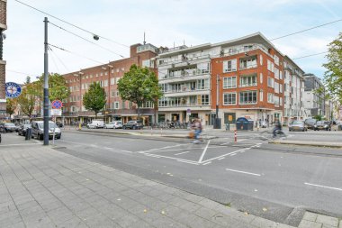 Kenarında arabaları olan bir şehir caddesi ve binaların, ağaçların ve çalıların önünde yürüyen insanlar.