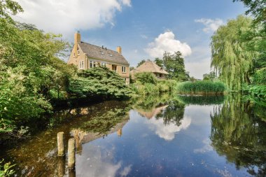 Arka planda ağaçlar ve su yüzeyine yansıyan mavi gökyüzü ile suların kenarında bir ev.