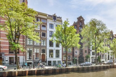 Amsterdam, Hollanda - 10 Nisan 2021: Bazı binalar ön kısımlarında ağaçlar ve arabaların park edildiği bir su kütlesi üzerindedir.