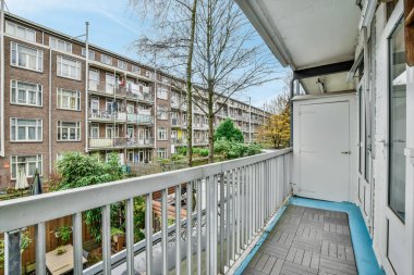 Arka planda ağaçlar ve binalar olan bir balkon. Bir apartmanın penceresinden sokağa bakıyor.