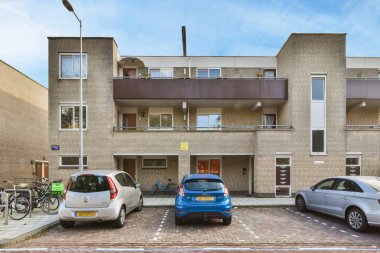 Tuğla bir binanın önüne park edilmiş iki araba diğer tarafta bir daire ve yanında da bir araba var.