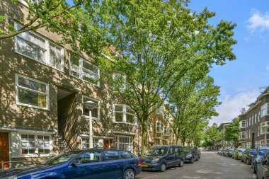 Amsterdam, Hollanda - 10 Nisan 2021: Sokağın her iki tarafında arabaların ve ağaçların park ettiği cadde apartmanlarla dolu.