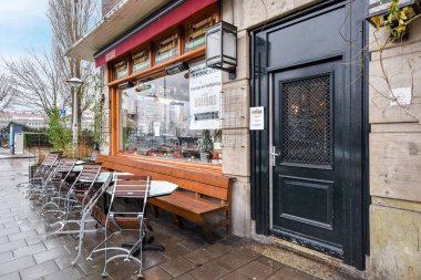 Amsterdam, Hollanda - 10 Nisan 2021: Fotoğrafın sağ tarafında açık kapısı olan bir binanın önünde bazı masa ve sandalyeler var.