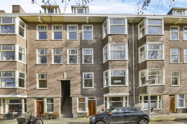 Bir apartmanın yanında iki araba park etmiş ve insanlar binanın önünde yürüyor.