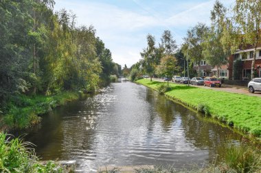 Amsterdam, Hollanda - 10 Nisan 2021: Her iki tarafında yemyeşil çimlerle çevrili evler ve ağaçlar olan bir bölgenin ortasındaki bir nehir.