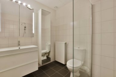 Beyaz bir banyo, siyah fayanslı döşeme ve duvarlarında fayanslar olan, ve köşesinde tuvalet olan.