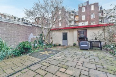 Duvarları tuğlalı ve yerde yeşil bitkileri olan bir avlu, köşede bir barbekü ızgarası var.