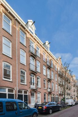 Beyaz camları ve bacakları olan tuğla bir binanın önündeki caddenin kenarına park etmiş birkaç araba.