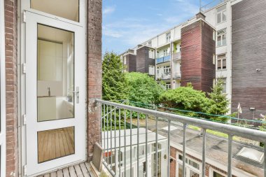 Fotoğrafın sol tarafında ahşap döşeme ve beyaz korkuluklu bir balkon var. Arka planda bir apartman var.