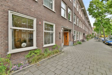 Kenara park edilmiş arabalarla tuğlalı bir cadde ve apartmanların önündeki kaldırımda ağaçlar var.