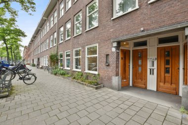 Beyaz pencereli ve kapılı kırmızı tuğlalı bir binanın önündeki caddenin kenarına parketmiş bisikletler.