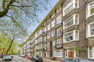 Apartman binalarının önünde caddenin iki yanına park etmiş arabaların ve ağaçların oluşturduğu bir cadde.