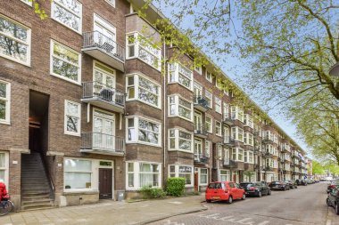 Birçok pencere ve balya olan bir apartmanın önüne park etmiş birkaç araba var.