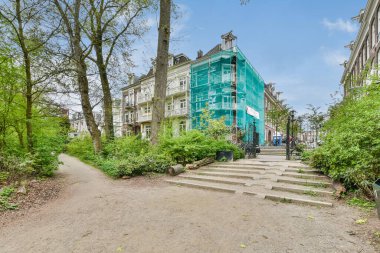 Ağaçların, çalıların ve merdivenlerin bulunduğu bir şehir alanı. Parlak maviyle boyanmış bir binaya çıkıyor.