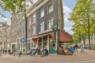 Amsterdam, Hollanda - 10 Nisan 2021: İnsanlar kaldırım boyunca ağaçlarla ve park edilmiş arabalarla dolu tuğlalı bir caddede bir restoranın önündeki masalarda oturuyorlar