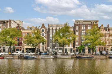 Amsterdam, Hollanda - 10 Nisan 2021: sudaki bazı tekneler ve nehrin diğer tarafındaki ağaçların arkasında kanalı kapladığı binalar