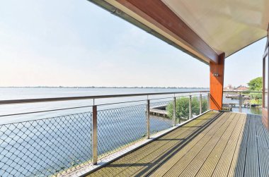 Balkondan suya bakan ahşap bir güverte. Bir tarafında turuncu bir kapı diğer tarafında da metal bir korkuluk var.