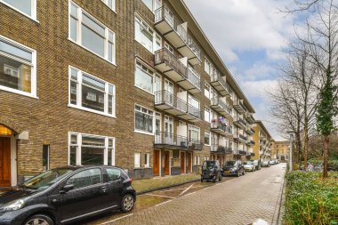 Bazı arabalar sokağın kenarına park edilmiş. Tuğla bir binanın önünde. Bir sürü pencere ve balya var.