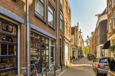 Amsterdam, Hollanda - 10 Nisan 2021: Tuğlalı binalar ve mavi gökyüzü olan bir kentsel alanda caddenin kenarına park edilmiş bazı arabalar
