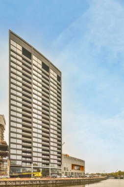 Amsterdam, Hollanda - 10 Nisan 2021: Önünde arabalar park edilmiş bir su kütlesi üzerinde bir bina ve etrafında yürüyen insanlar