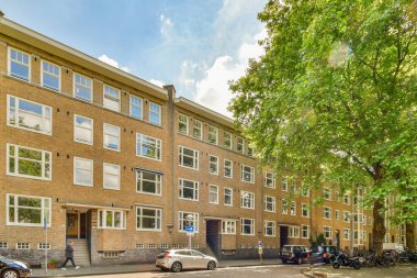 Bir apartman dairesi. Arabalar hemen yanına park edilmiş ve ağaçlar yol kenarında dizilmiş.