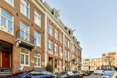 Bazı arabalar sokağın kenarına park etmiş. Tuğla bir binanın önünde. Bir sürü pencere ve balya var.