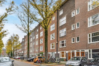 Amsterdam, Hollanda - 10 Nisan 2021: Caddenin kenarına park etmiş arabalar sokak boyunca ağaçlarla kaplı tuğla apartmanların önünde.