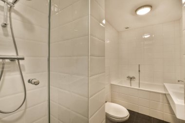 Küvetin köşesindeki tuvaletin yanında siyah döşemeli, beyaz fayanslı modern bir banyo var.