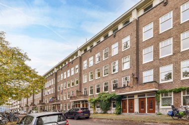 Yolun kenarına arabaların park ettiği tuğladan bir apartman dairesi ve yol kenarında ağaçlar.