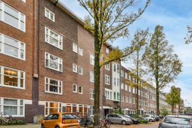 Amsterdam, Hollanda - 10 Nisan 2021: birçok penceresi ve ağacı olan bir tuğla binanın önündeki caddenin kenarına park edilmiş arabalar