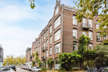 Amsterdam, Hollanda - 10 Nisan 2021: Ağaçlarla çevrili bir apartmanın önündeki kaldırımda birçok park etmiş araba ve bisikletin bulunduğu bir şehir caddesi.