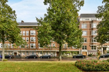 Amsterdam, Hollanda - 10 Nisan 2021: Büyük tuğla bir binanın önüne park edilmiş bazı arabalar ve yerde bir sürü yeşil yaprak var.