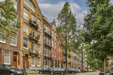 Amsterdam, Hollanda - 10 Nisan 2021: Bir caddenin kenarına park etmiş bazı arabalar yüksek tuğlalı binalar ve bir sürü yeşil ağaçlar.
