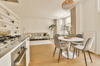 Beyaz duvarları, ahşap döşemeleri ve sokağa bakan büyük pencereleri olan bir evde mutfak ve yemek odası.